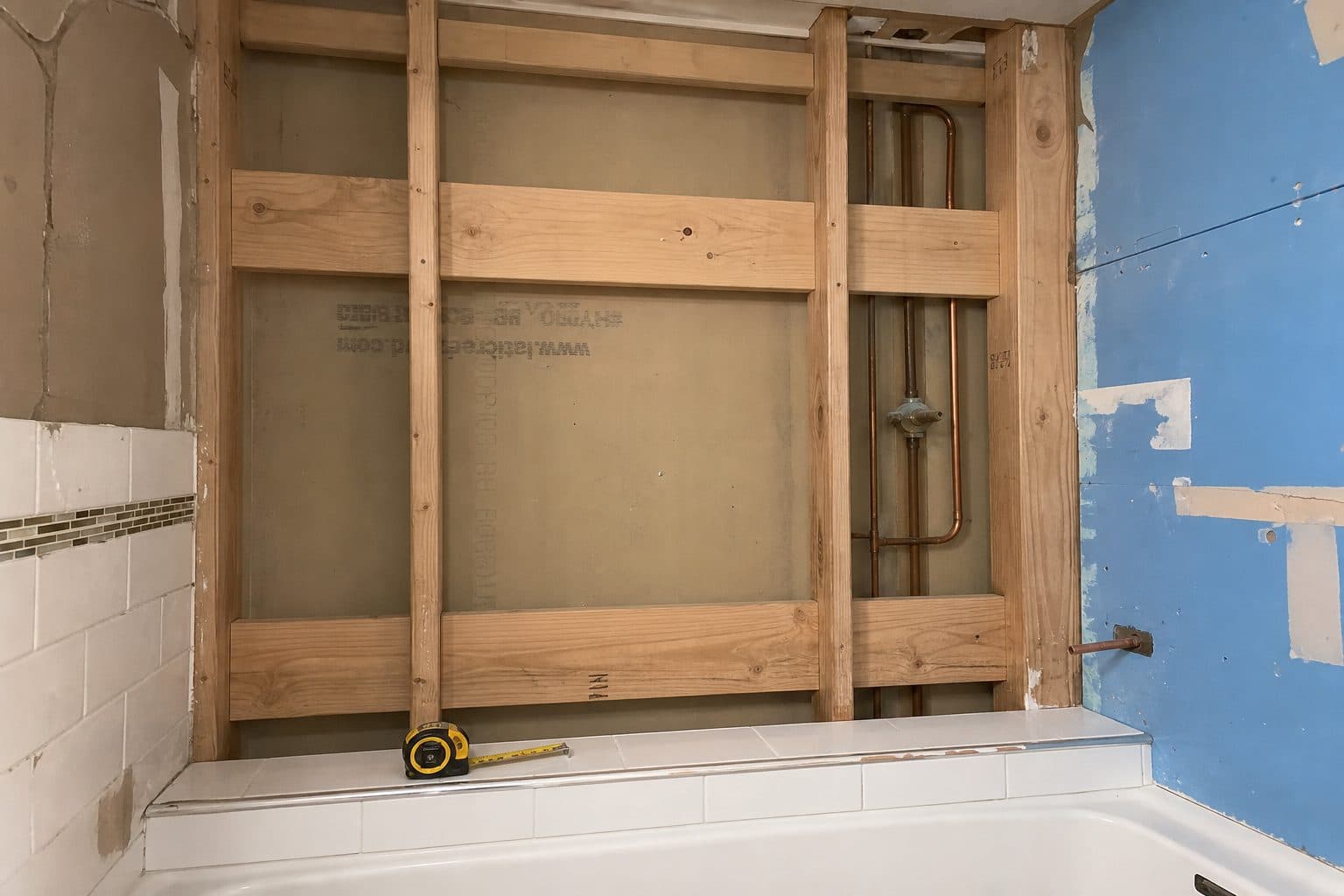 Open bathroom wall showing wood blocking for grab bars, tape measure on the tile lip.